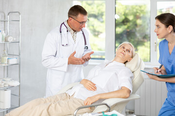 Obraz premium Elderly woman sitting in chair at clinic reception and carefully answers male doctor questions about his well-being, health complaints, nurse records data in client medical record