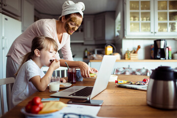 Young mother and daughter having breakfast and using a laptop in the morning in the kitchen