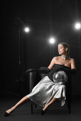 Fototapeta na wymiar Stylish young woman with disco ball sitting in armchair on dark background