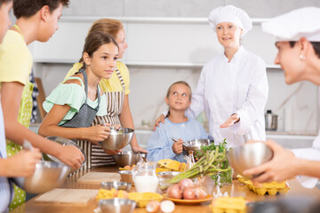 Interested teen girl taking part in cooking classes for children, mixing sauce in bowl with whisk, carefully listening to advices of professional chef instructor