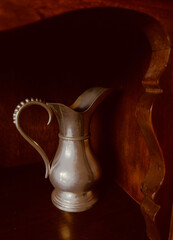 Old pewter pitcher closeup on shelf. S curves & shadows with text space in dark area is available. Wood furniture with rich tones is vintage.