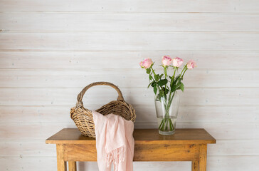 Close up of pale pink roses in glass vase on oak side table with wicker basket and scarf against rustic wood panelled wall (selective focus) - Powered by Adobe