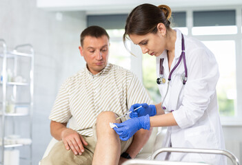 Young female doctor performs intra-articular injection treatment of knee bursitis to middle-aged man patient