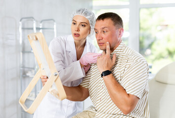 Obraz premium Portrait of man sitting on couch and looking at mirror during appointment with doctor at cosmetology clinic