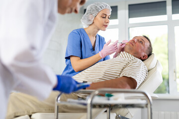 Obraz premium Experienced female cosmetologist working with male client in clinic of aesthetic medicine, performing minimally invasive procedure using filler injections to correct lips contour