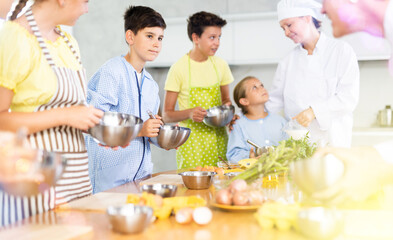 Interested tween boy taking part in cooking classes for children, mixing sauce in bowl with whisk, carefully listening to advice of professional chef instructor