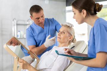 Fototapeta premium Interested elderly woman sitting on examination couch and looking at mirror while professional cosmetologist with female assistant planning and explaining future rejuvenating facial procedure