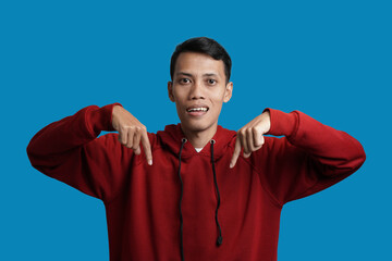 Handsome cheerful young Asian man wearing red hoodie pointing at copy space place, showing place for your advertisement isolated over blue background.