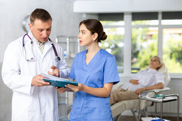 Experienced male doctor advising young female colleague about diagnosis of patient while reviewing case history in clinic..