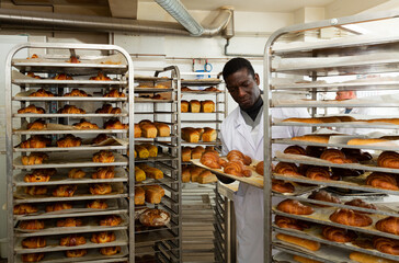 Skilled African American baker arranging trays with freshly baked bakery products on trolley..