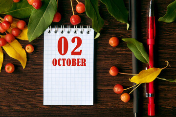 calendar date on wooden dark desktop background with autumn leaves and small apples.  October 2 is...