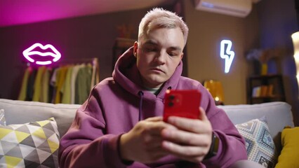 Fair-haired guy with phone on sofa in living room-studio neon lights in background. Expression of negative emotions anger disappointment frustration. Writes messages in anger. Virtual modern lifestyle