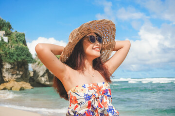 A happy young Asian woman wearing beach hat is posing to the camera with her hands open, expressing freedom and carefree holiday at the beach in Gunungkidul, Indonesia