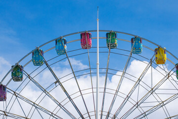  a photo of a rainbow ferris wheel that stands out against a blue sky with white clouds.
