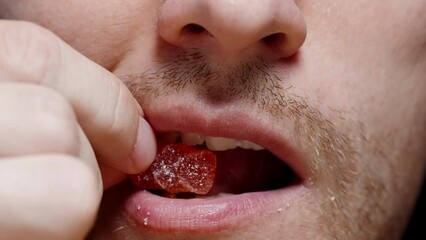 In a close-up shot, a bearded man places a teddy bear-shaped marmalade candy into his mouth.