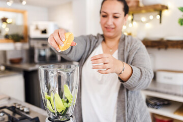 Latin woman overweight preparing detox or green juice in kitchen at home in Mexico Latin America, hispanic female plus size - obrazy, fototapety, plakaty