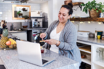 young latin woman overweight using laptop while having breakfast at home kitchen in Mexico Latin...