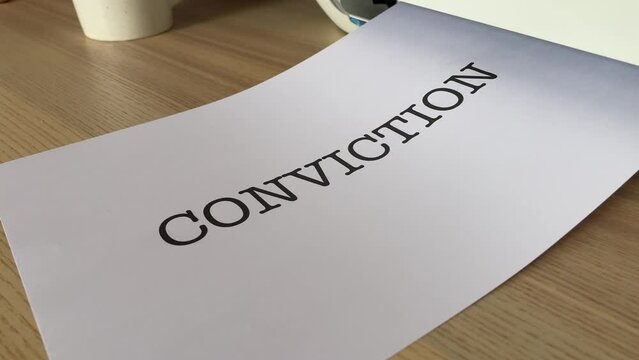 “Conviction” black text word on paper sheet A4 document sliding out of printer scanner on wooden table close up view