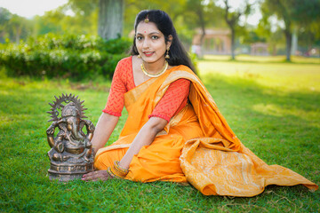 Happy young indian woman wearing saree sitting along with lord Ganesha idol outdoor at park...