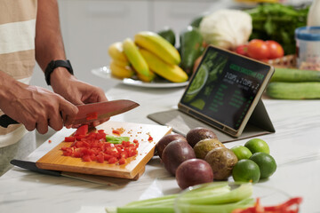 Hands of young modern man with sharp knife chopping fresh red capsicum on wooden board while...