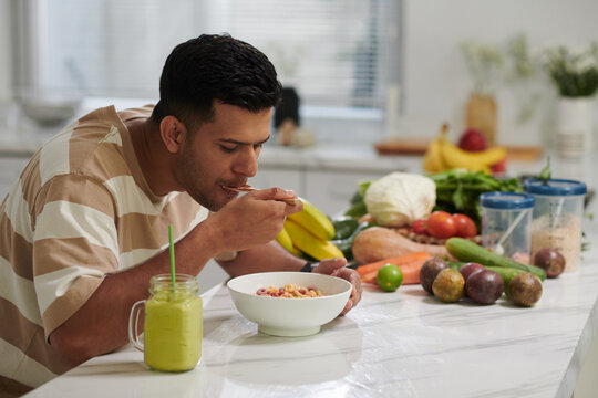 Young hungry man eating muesli from bowl and having fresh juice or smoothie while sitting by kitchen table with fruits and vegetables