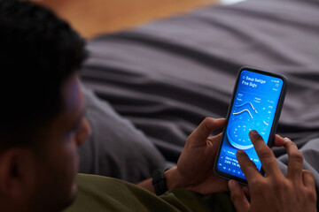 Focus on hands of young man choosing time settings in smartphone while lying on bed in the evening before sleep and switching on alarm