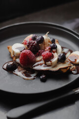 Vertical photo of dark food photography: tasty breakfast of crepes, made with wheat flour, decorated with dulce de leche and chocolate syrup, with delicious red berries, on a black plate