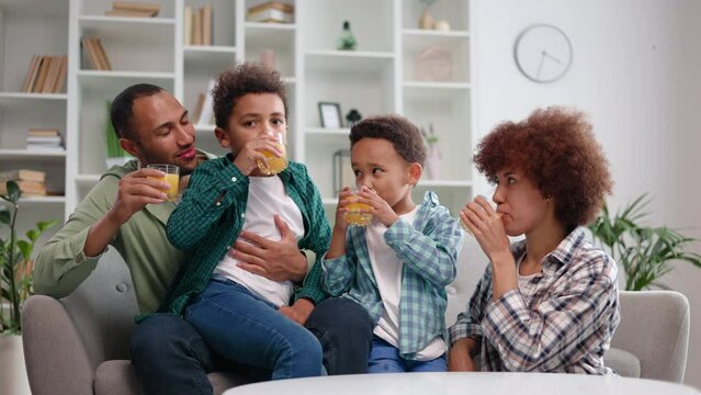 Happy african american adults and chidren in everyday clothes having sip of beverages during mealtime at home. Smiling parents and playful sons celebrating family traditions with fresh juice