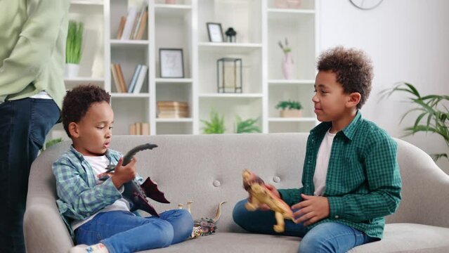 Two african american male kids dressed in casual clothes resting on grey couch and playing with toys in apartment. Cute little boys entertaining together while father talking on smartphone