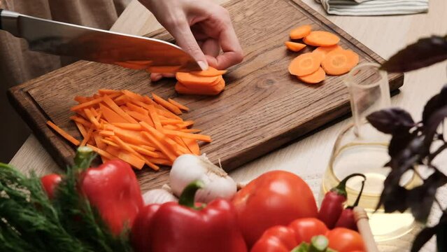 Close-up of a woman cutting fresh carrot with a knife in the kitchen. Preparing vegetable salad. Housewife chopping carrot with knife on wooden cutting board