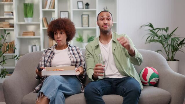 Emotional Male Sport Fan Sitting On Couch Together With Girlfriend And Watching Football Game With Bottle Of Drink In Hands. Biracial Lady Holding Pizza On Knees Being Uninterested And Bored