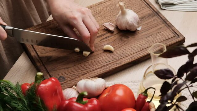 Close-up of a woman cutting fresh garlic with a knife in the kitchen. Preparing vegetable salad. Housewife chopping garlic with knife on wooden cutting board