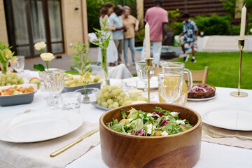 Focus on wooden bowl with fresh homemade vegetarian salad standing on served table among plates, forks, fruit, candles, glasses and flowers