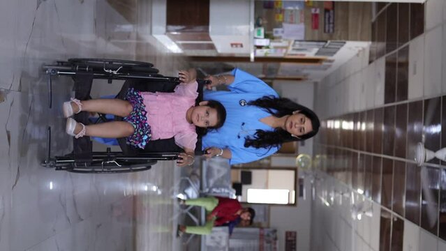 Doctor bringing the wheel chair young girl child patient in bright corridor. Front view of mature woman doctor in blue coat taking care of young girl on wheelchair. Having a stroll in the corridor.