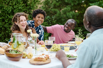 Cheerful young couple and their cute son having fun by served table and enjoying outdoor dinner with mature parents at backyard of country house