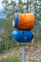 Yellow snow cannon on the edge of a hill overlooking a valley in summer or warm winter, green mountain peaks. Ski slope without snow in summer or during warm winter, greenhouse effect concept.