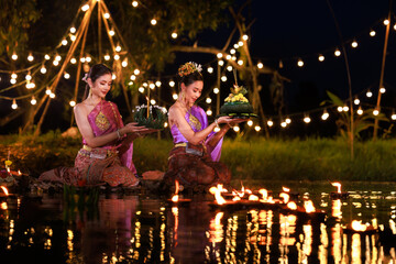 Two thai woman holding a krathong sitting on a raft by the river, Asian women in traditional Thai...