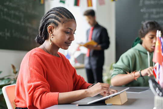 African American student using digital tablet while sitting at table with her classmates during lesson
