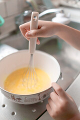 卵をかき混ぜている子供の手（小学生低学年）。ホットケーキ、お菓子作り、卵料理。泡立て器とボール