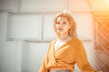 A middle-aged woman looks like a good blonde with curly beautiful hair and makeup on the background of the building. She is wearing a yellow sweater.