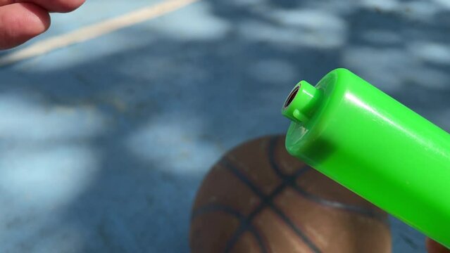Male hand tighten green inflatable ball pump air needle outdoor sunny day at basketball court with blurred ball on blue ground floor. Sport and recreation concept