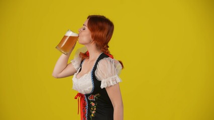 Medium yellow background isolated side-view shot of a young German woman, waitress, wearing a traditional costume, taking a sip, gulp of beer with pleasure.