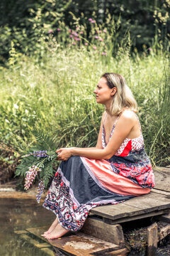 A beautiful young woman with blond hair in a sundress on the shore of a lake with a bouquet of wildflowers. National Russian clothes . Country life