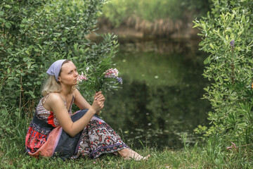 A beautiful young woman with blond hair in a sundress on the shore of a lake with a bouquet of wildflowers. National Russian clothes . Country life