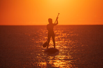 Fototapeta premium SUP. Stand up shovel. Adventurous girl on the paddle board rows during a bright and vibrant sunset.Silhouette of woman paddleboarding at sunset. Canakkale, Türkiye.
