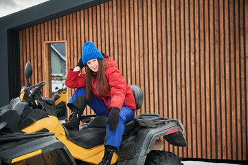 Portrait of beautiful woman posing on offroad four-wheeler ATV with wooden house on background. Concept of active leisure and winter activities.