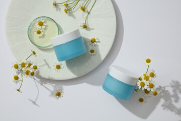 Flat lay of two unlabeled blue jars arranged with glass petri dish and flowers. Chamomilla (Matricaria chamomilla) is very useful in cosmetic production