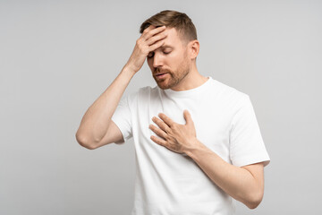 Unhealthy overwhelmed man with unhappy expression covered forehead hand feel bad, chest pain, fever. Unwell haggard male pressed hand to heart in horror from terrible news, isolated studio background.