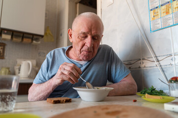 Elderly senior man eating soup with bread and green onion at home in the kitchen. Happy retirement,...
