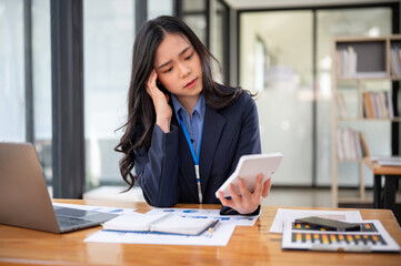 A thoughtful and stressed Asian businesswoman is using a calculator and working at her desk.